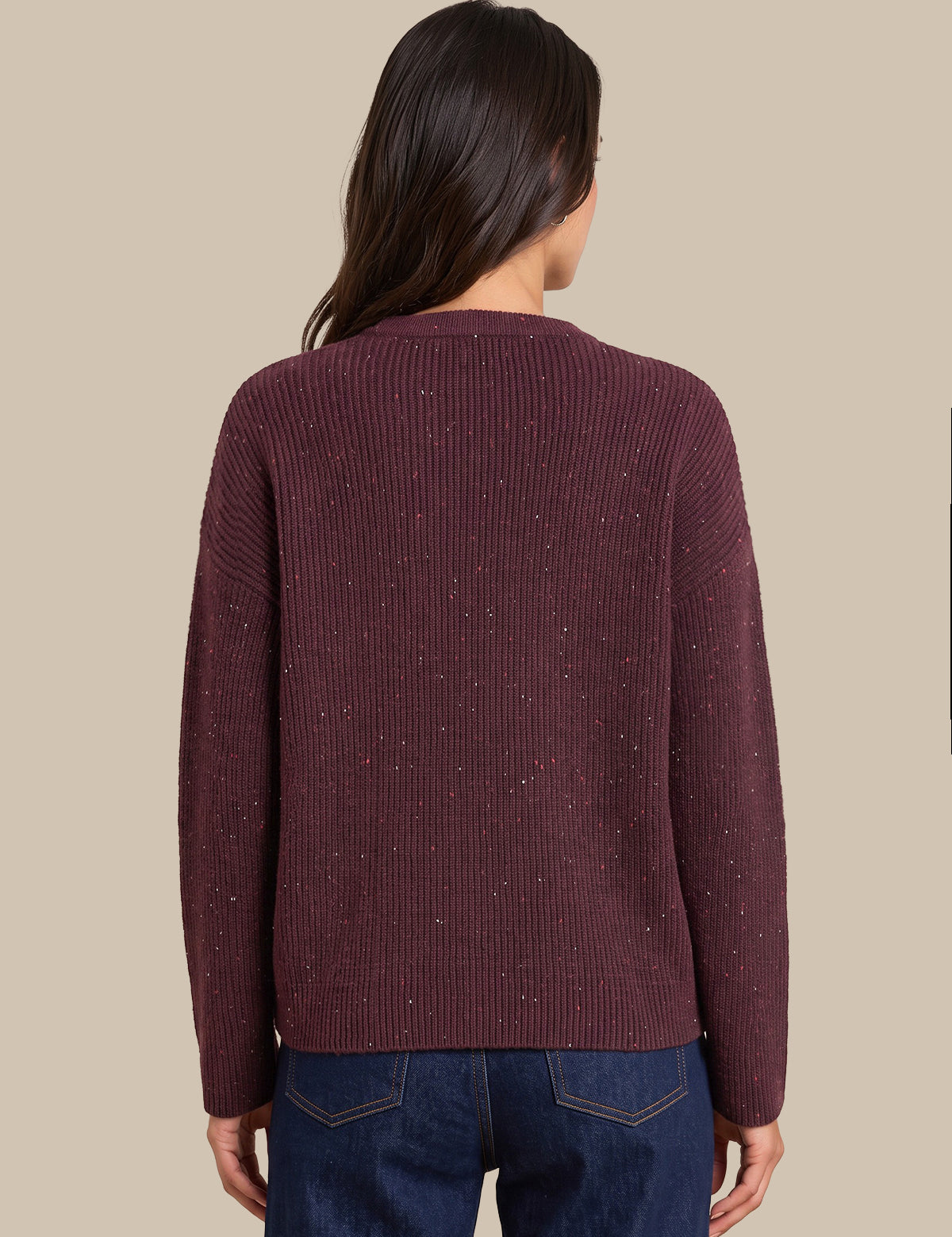 A woman viewed from the back wearing a ribbed knit sweater in a deep burgundy color with subtle speckled flecks, paired with dark blue jeans, against a solid beige background.
