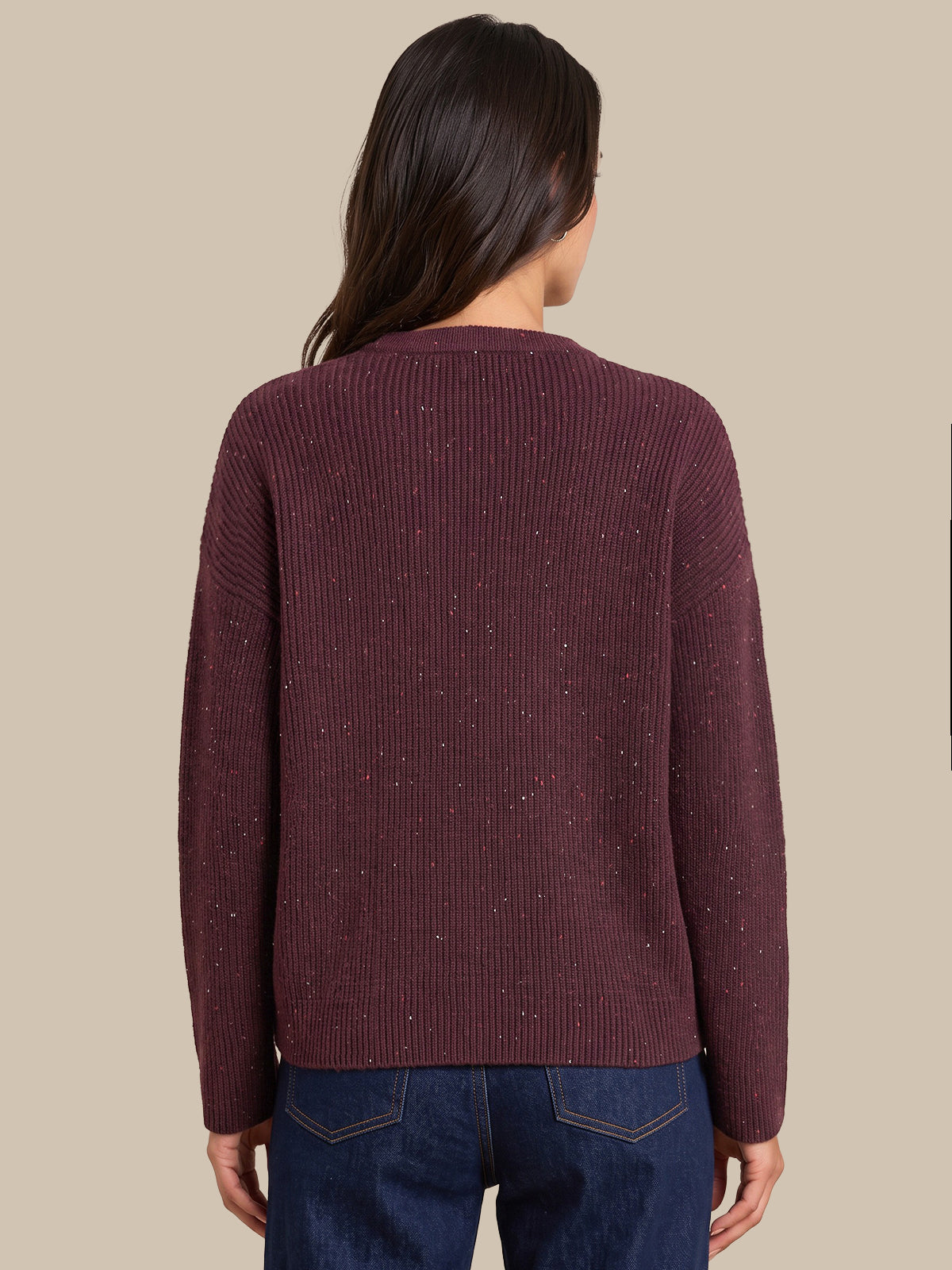 A woman viewed from the back wearing a ribbed knit sweater in a deep burgundy color with subtle speckled flecks, paired with dark blue jeans, against a solid beige background.