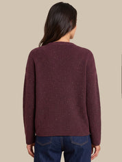 A woman viewed from the back wearing a ribbed knit sweater in a deep burgundy color with subtle speckled flecks, paired with dark blue jeans, against a solid beige background.