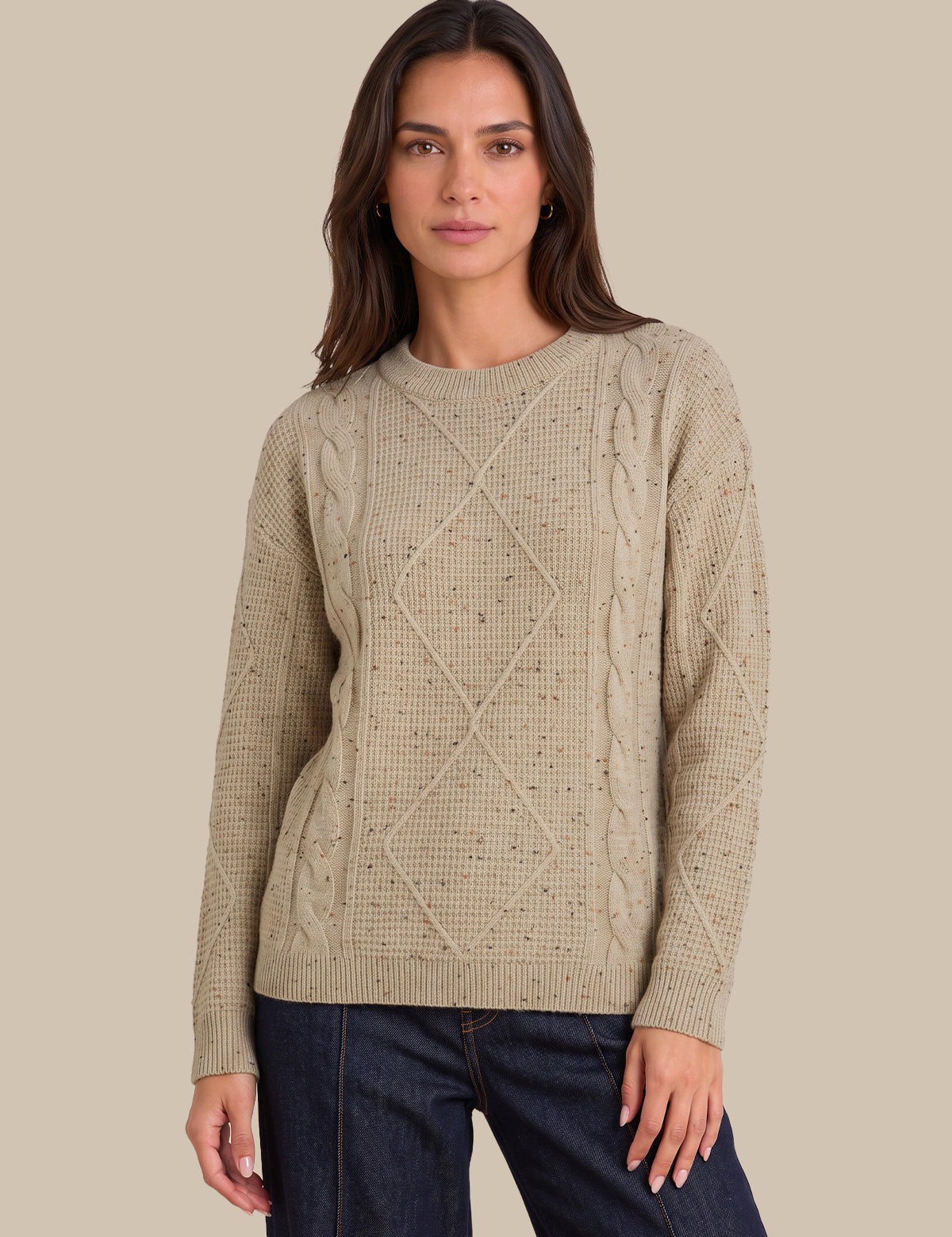 A woman modeling a beige cable-knit crewneck sweater with textured knit details, paired with dark blue jeans, against a solid beige background.