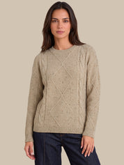 A woman modeling a beige cable-knit crewneck sweater with textured knit details, paired with dark blue jeans, against a solid beige background.