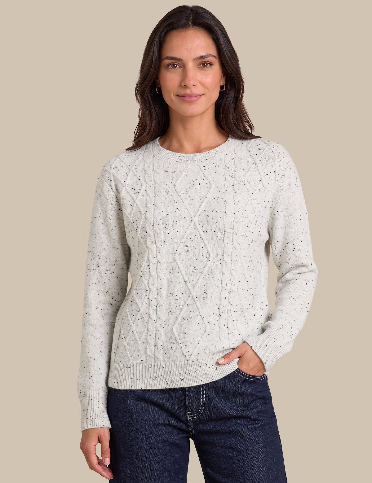 A woman modeling a light cream cable-knit sweater with subtle dark speckles, paired with dark blue jeans, against a solid beige background.