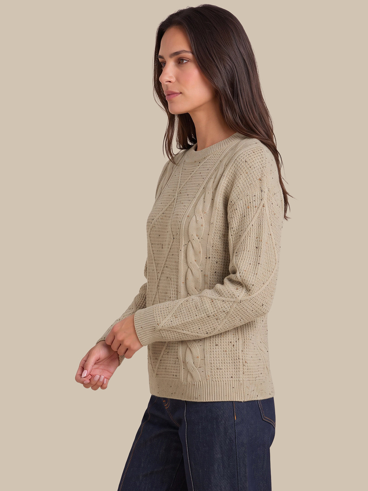 A woman wearing a beige cable-knit crewneck sweater and dark blue jeans, shown in profile against a solid beige background.