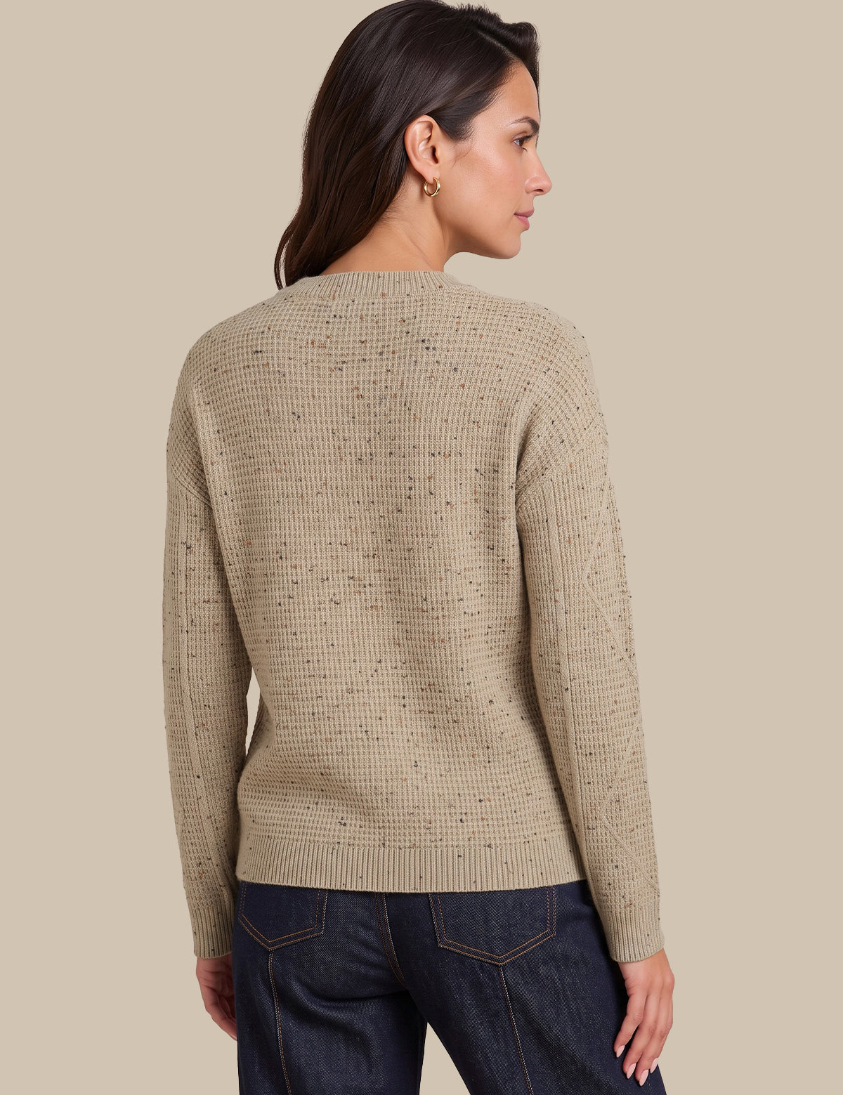 A woman shown from the back wearing a beige textured knit sweater with subtle dark speckles and dark blue jeans, set against a solid beige background.