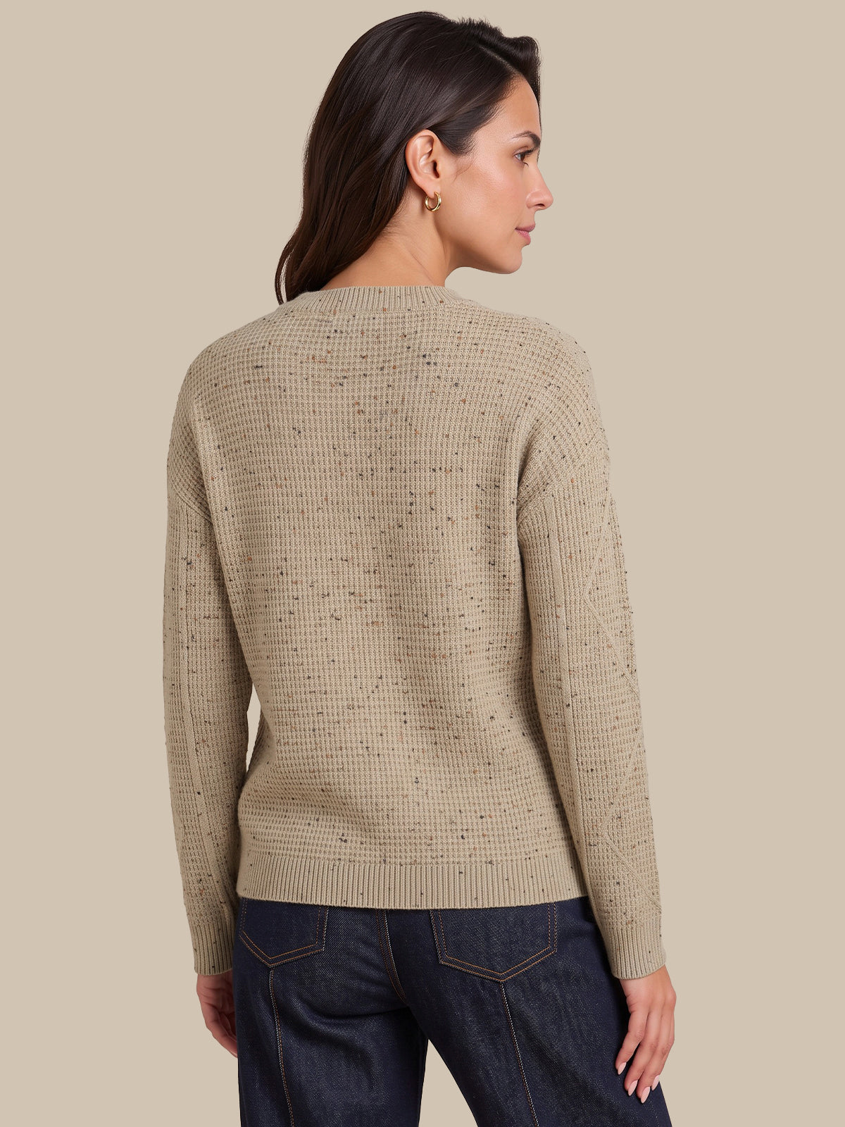 A woman shown from the back wearing a beige textured knit sweater with subtle dark speckles and dark blue jeans, set against a solid beige background.