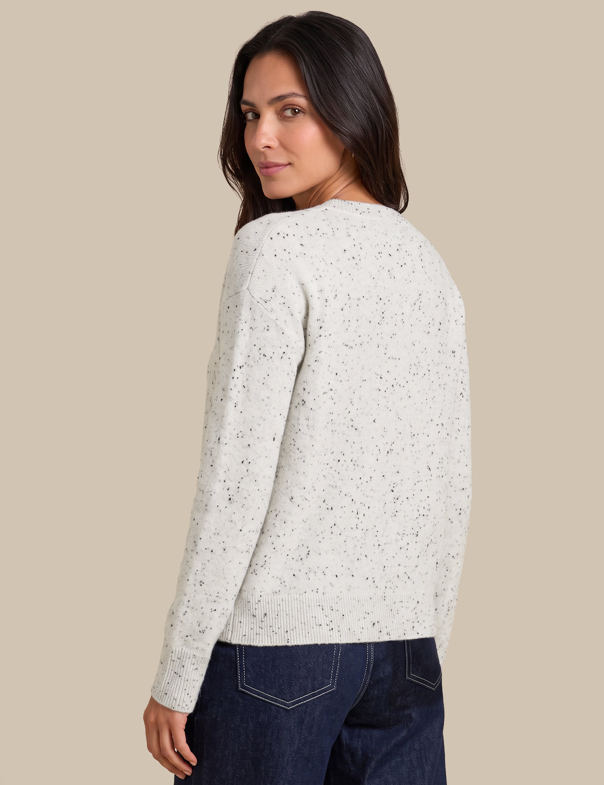 A woman wearing a cream-colored knit sweater with tiny black speckles, shown from a three-quarters back view against a solid beige background.