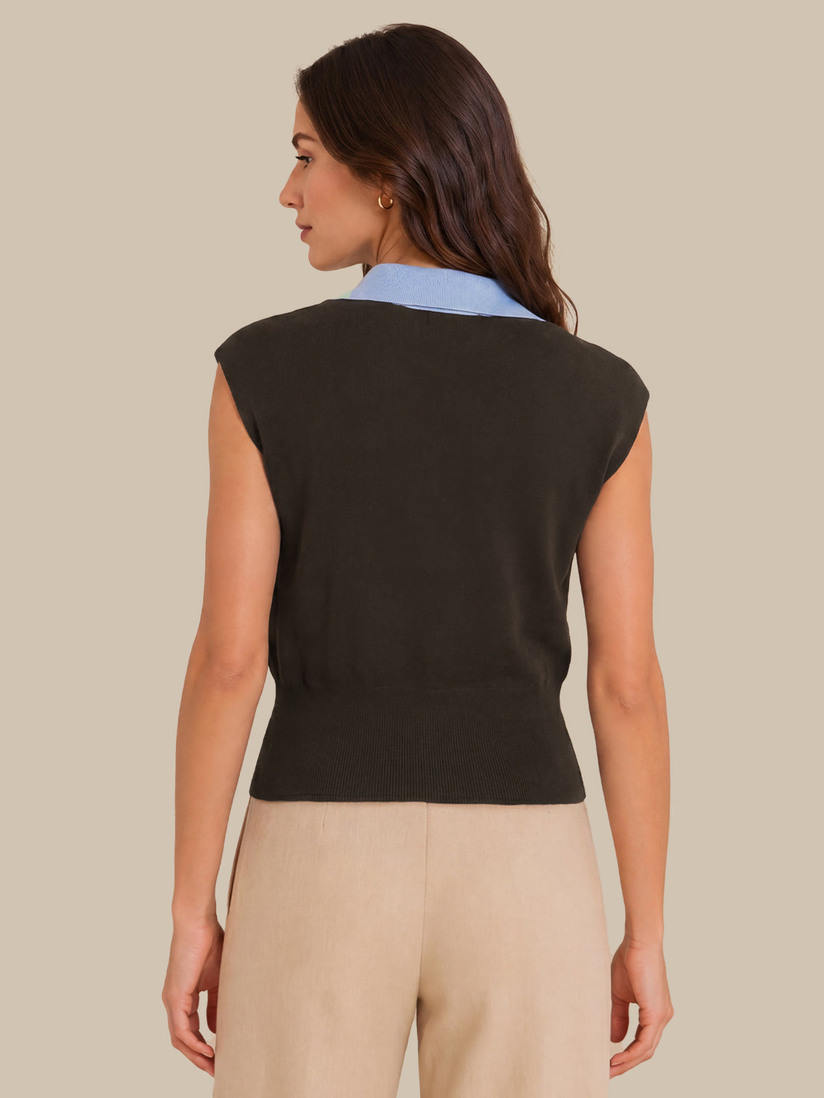 A woman shown from the back wearing a sleeveless black knit vest over a light blue collared shirt, paired with beige pants, against a solid beige background.