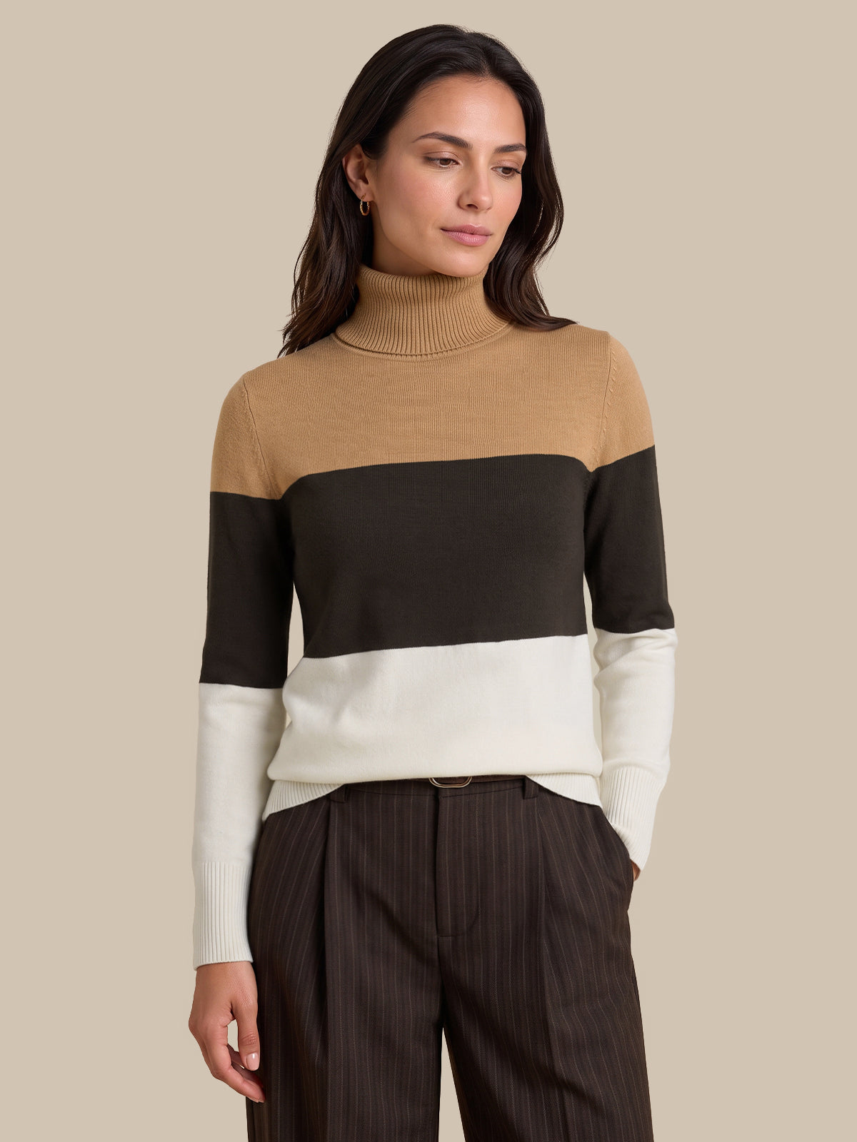 A woman wearing a color-blocked knit turtleneck sweater with beige, black, and white horizontal bands, paired with dark brown pinstriped trousers, against a solid beige background.