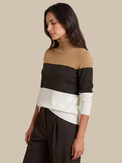 A woman wearing a color-blocked knit turtleneck sweater with horizontal stripes in camel, black, and white, paired with dark pinstriped trousers, set against a solid beige background.
