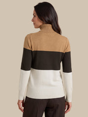 A woman shown from the back wearing a color-blocked ribbed turtleneck sweater with horizontal stripes in tan, black, and off-white, paired with dark brown pinstriped trousers, against a solid beige background.