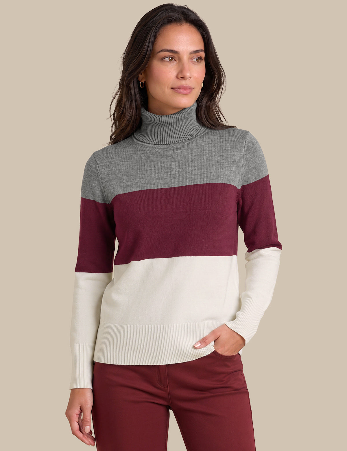 A woman wearing a color-blocked turtleneck sweater with gray, burgundy, and cream horizontal stripes, paired with burgundy pants, against a solid beige background.