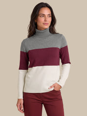 A woman wearing a color-blocked turtleneck sweater with gray, burgundy, and cream horizontal stripes, paired with burgundy pants, against a solid beige background.
