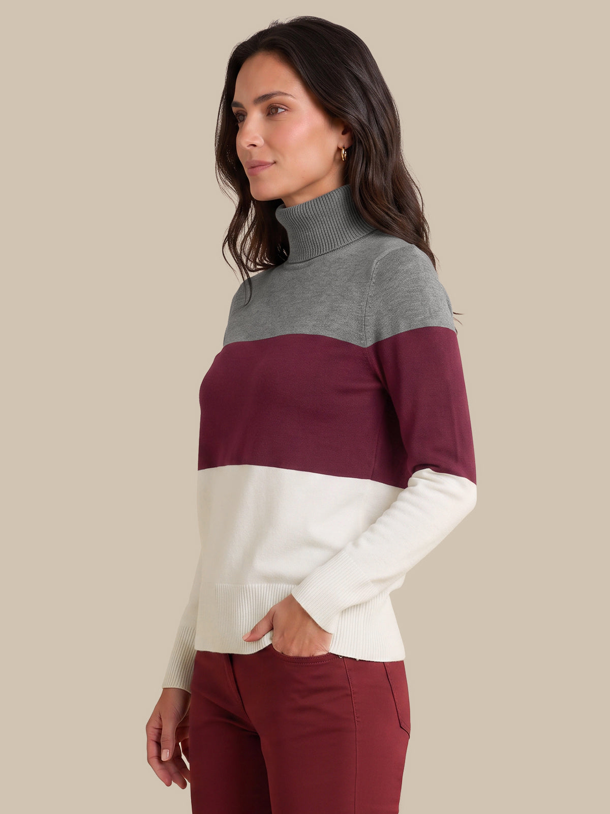 A woman wearing a color-blocked turtleneck sweater with gray, burgundy, and cream horizontal stripes, paired with burgundy pants, set against a solid beige background.