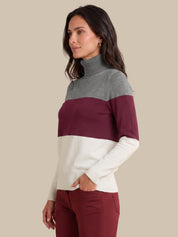 A woman wearing a color-blocked turtleneck sweater with gray, burgundy, and cream horizontal stripes, paired with burgundy pants, set against a solid beige background.