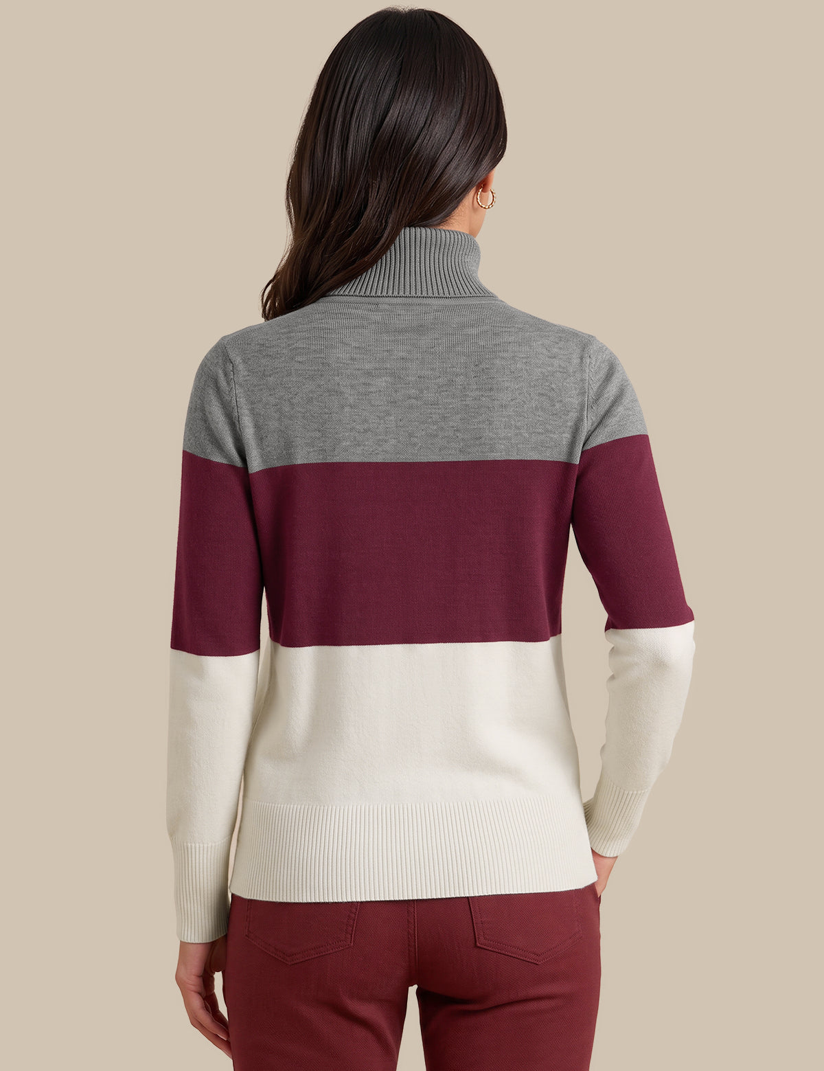 A woman viewed from the back wearing a color-blocked turtleneck sweater with a heather gray upper section, a wide burgundy middle band, and an off-white lower section, paired with burgundy pants against a solid beige background.