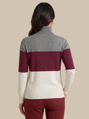 A woman viewed from the back wearing a color-blocked turtleneck sweater with a heather gray upper section, a wide burgundy middle band, and an off-white lower section, paired with burgundy pants against a solid beige background.