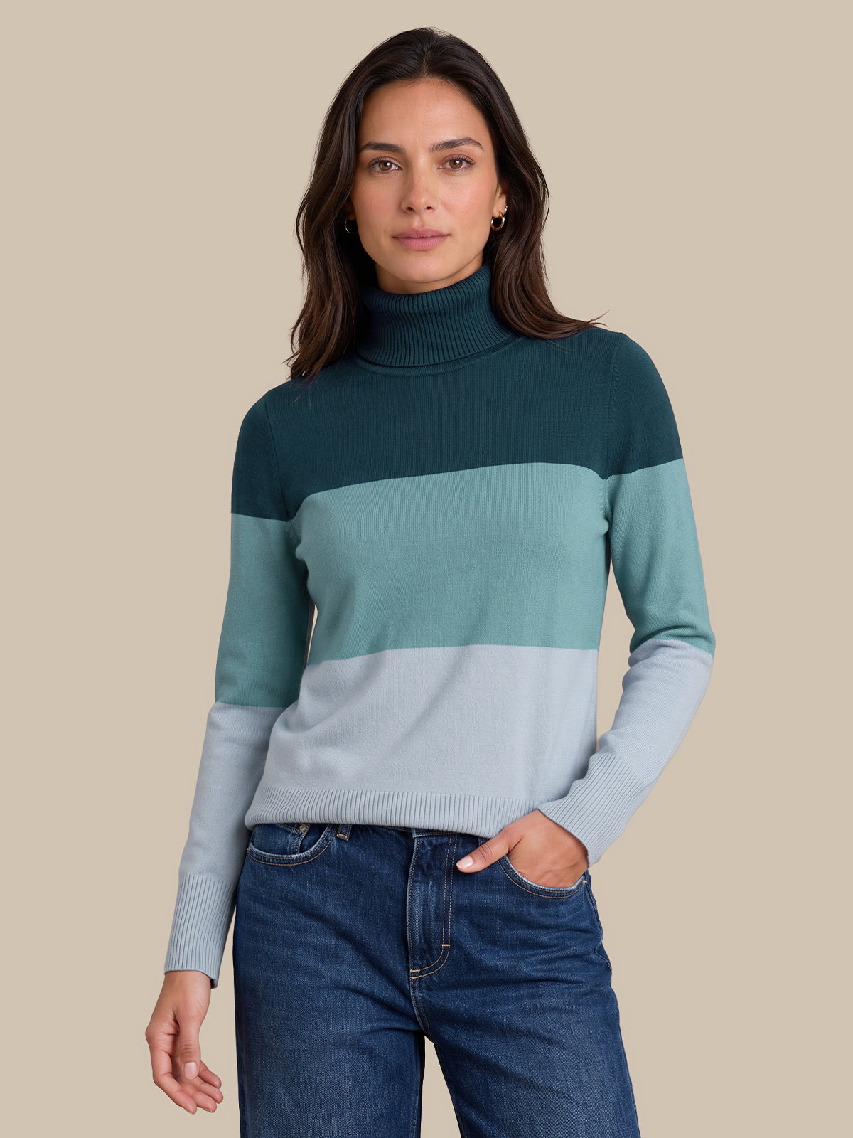A woman wearing a color-blocked turtleneck sweater with dark teal, mint green, and light gray horizontal stripes, paired with dark blue jeans, against a solid beige background.