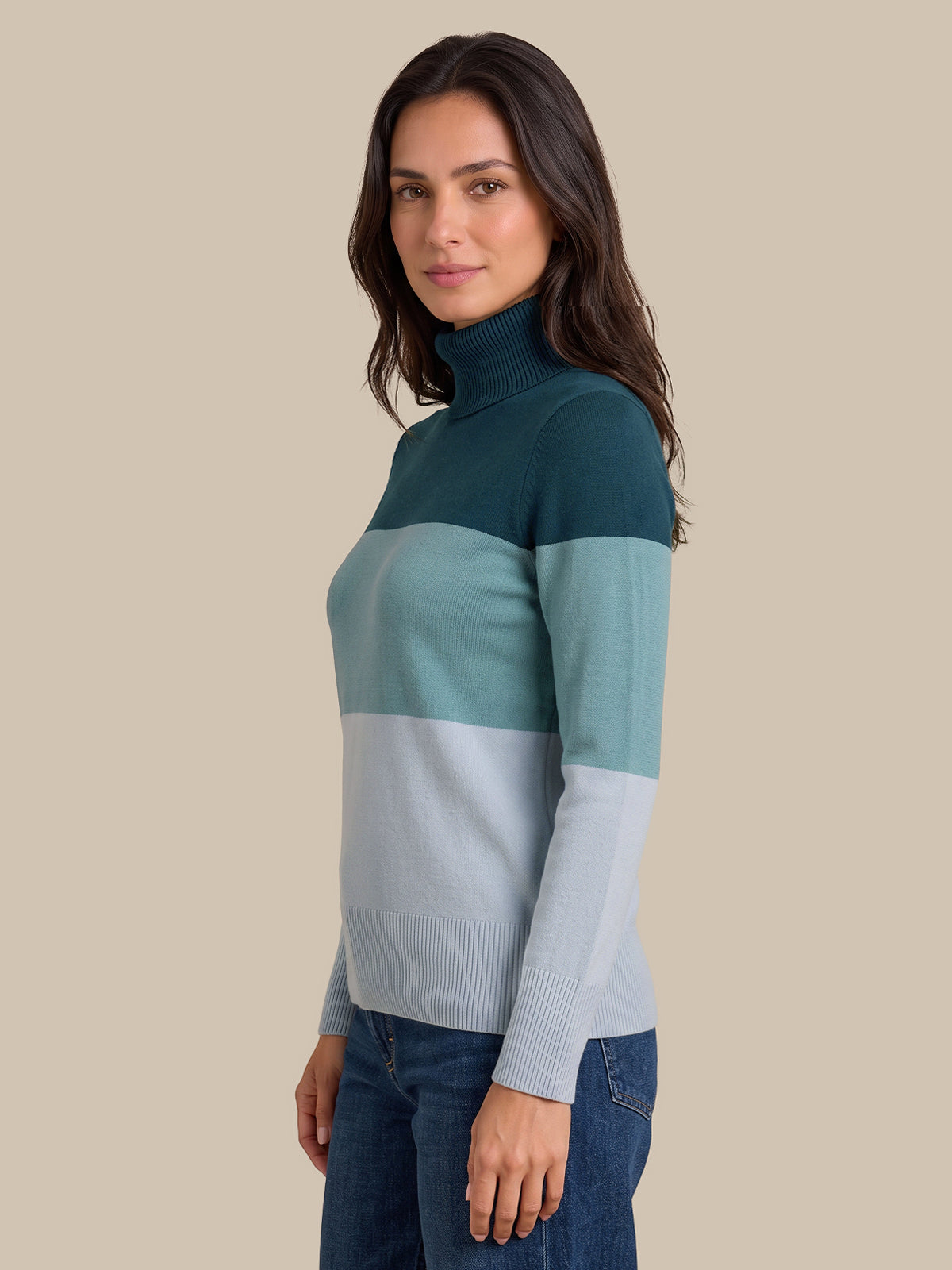 A woman wearing a colorblock turtleneck sweater with three horizontal color blocks: dark teal, light teal, and pale blue-gray, paired with dark blue jeans, set against a solid beige background.