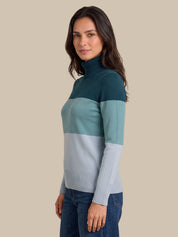 A woman wearing a colorblock turtleneck sweater with three horizontal color blocks: dark teal, light teal, and pale blue-gray, paired with dark blue jeans, set against a solid beige background.