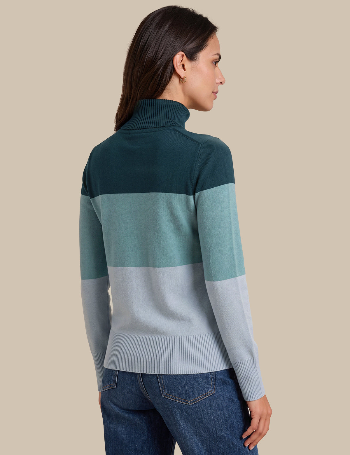 A woman shown from the back wearing a color-blocked rib-knit mock neck sweater with dark teal, mint green, and light gray horizontal stripes, paired with blue jeans, against a solid beige background.