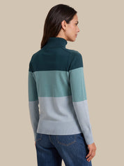 A woman shown from the back wearing a color-blocked rib-knit mock neck sweater with dark teal, mint green, and light gray horizontal stripes, paired with blue jeans, against a solid beige background.