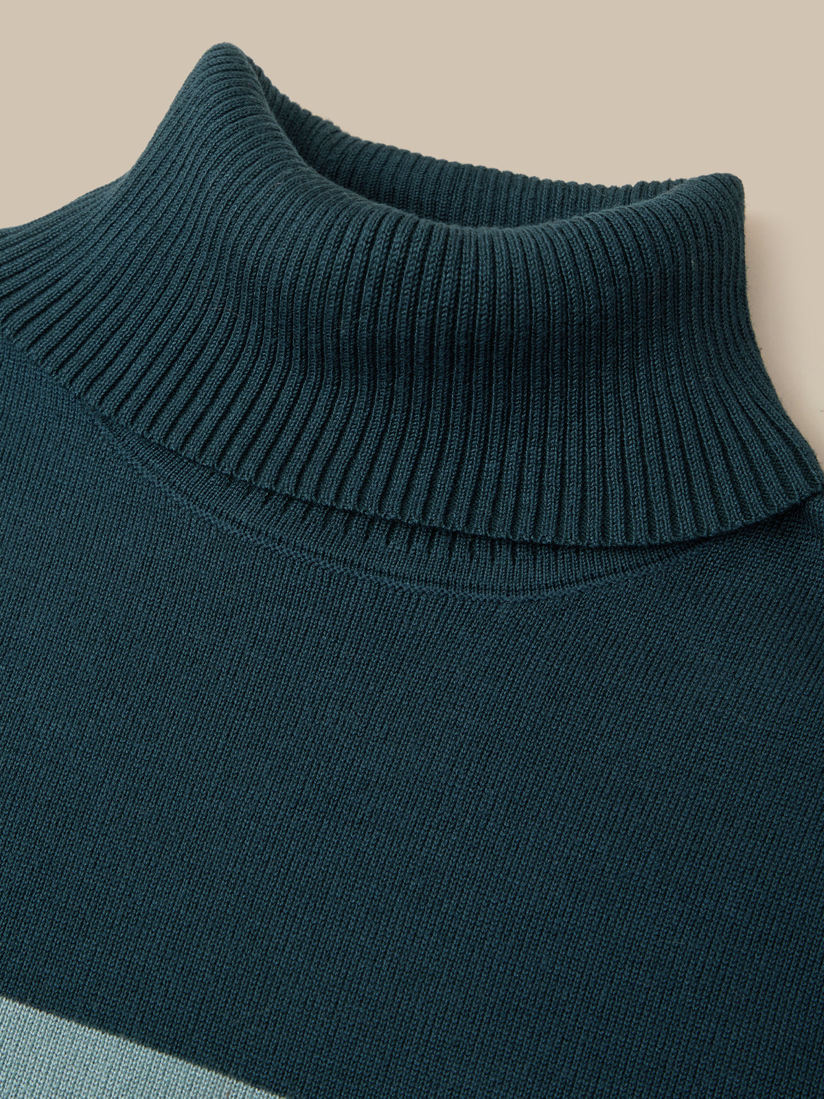 Close-up of a ribbed knit turtleneck sweater in a deep teal blue-green color, showing the texture of the collar and upper chest against a warm beige background.