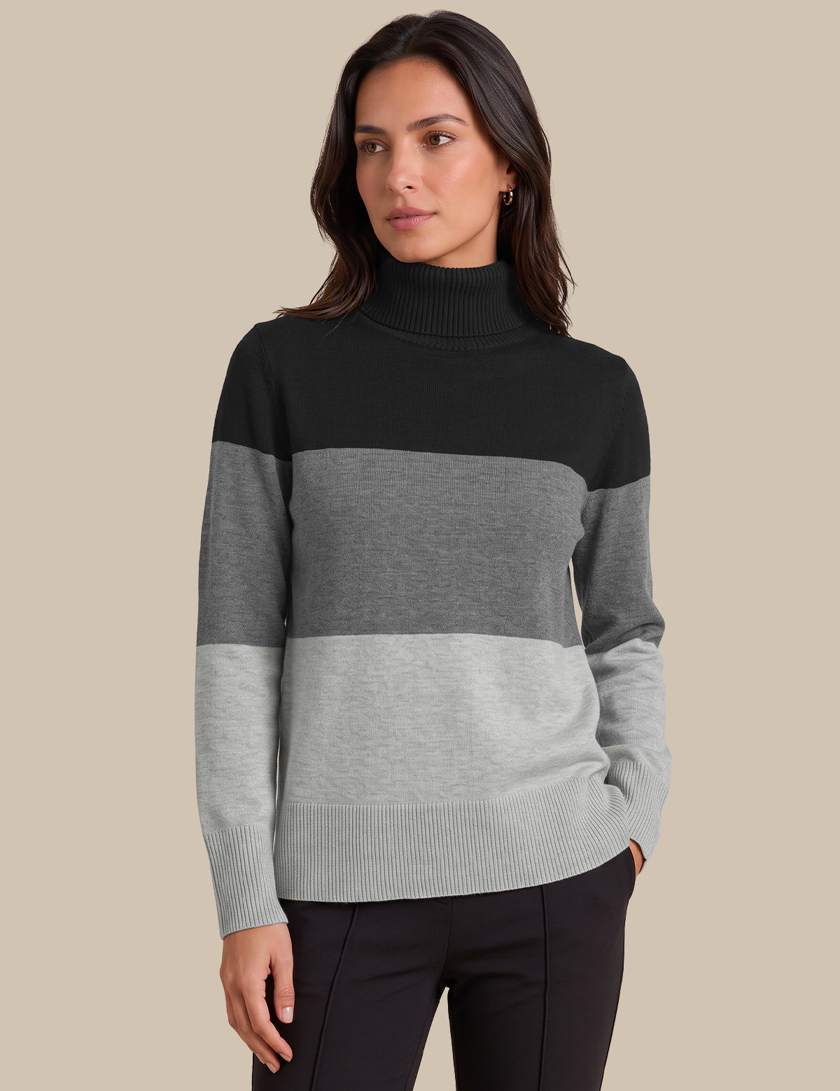 A woman modeling a colorblock mock neck sweater with horizontal stripes in black, medium gray, and light gray, paired with black pants against a solid beige background.