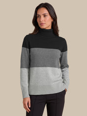 A woman modeling a colorblock mock neck sweater with horizontal stripes in black, medium gray, and light gray, paired with black pants against a solid beige background.