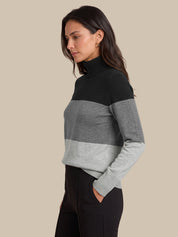 A woman wearing a colorblock mock neck sweater with black, medium gray, and light gray horizontal stripes, paired with black pants, set against a solid beige background.