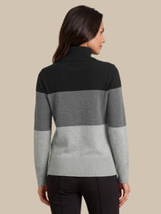 A woman shown from the back wearing a color-blocked knit mock-neck sweater with horizontal stripes in black, medium gray, and light gray, paired with black pants, against a solid beige background.