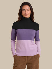 A woman wearing a color-blocked knit turtleneck sweater with a black upper section, a wide medium purple band, and a lighter lavender lower section, paired with brown corduroy pants against a solid beige background.