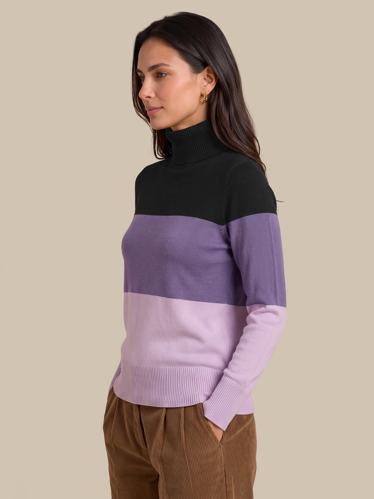 A woman wearing a color-blocked knit turtleneck sweater with a black yoke, a wide lavender band, and a light lavender lower section, paired with brown corduroy pants, set against a solid beige background.