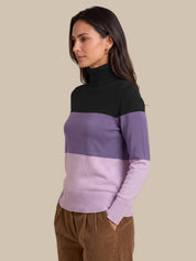 A woman wearing a color-blocked knit turtleneck sweater with a black yoke, a wide lavender band, and a light lavender lower section, paired with brown corduroy pants, set against a solid beige background.