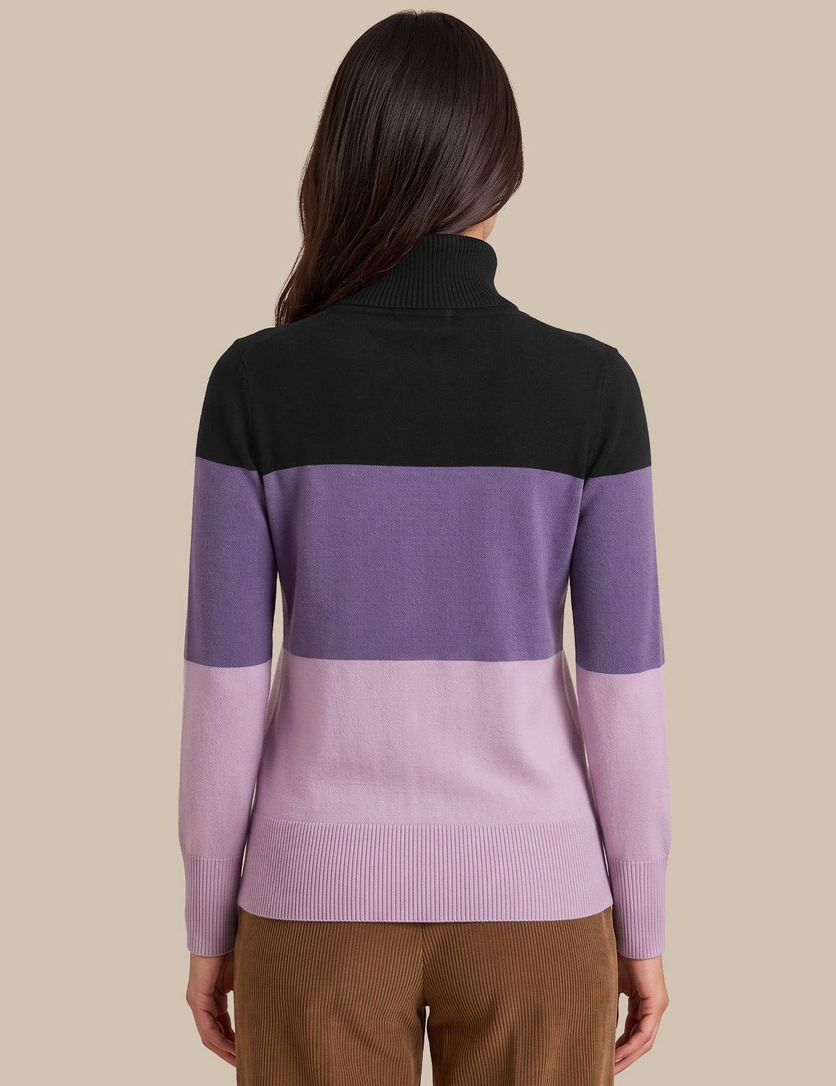 A woman shown from the back wearing a color-blocked knit sweater with a black yoke, a wide lavender-purple band, and a lighter pastel purple lower section and sleeves, paired with brown pants against a solid beige background.