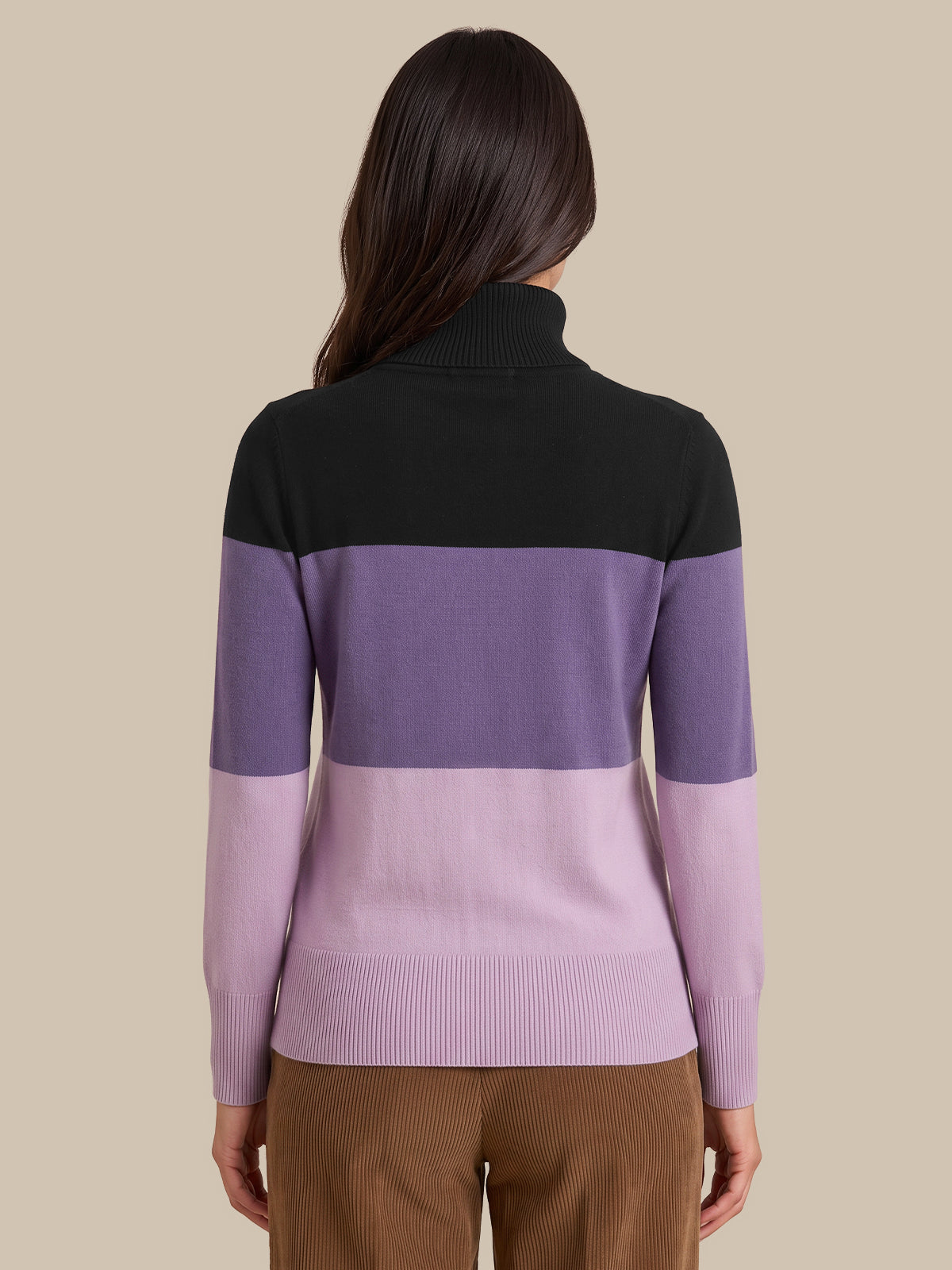 A woman shown from the back wearing a color-blocked knit sweater with a black yoke, a wide lavender-purple band, and a lighter pastel purple lower section and sleeves, paired with brown pants against a solid beige background.