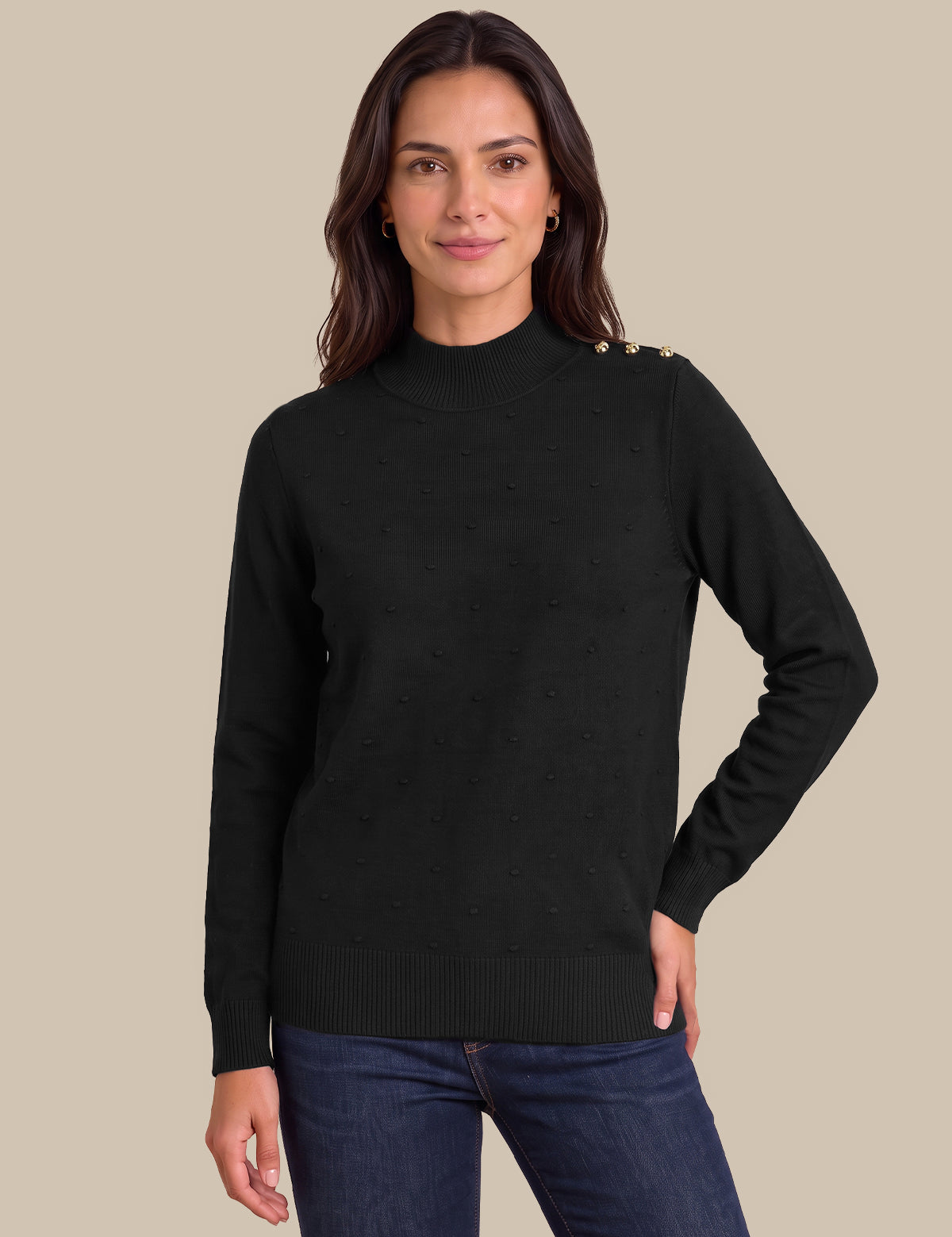 A woman wearing a black mock-neck knit sweater with gold button details on the shoulder, paired with dark blue jeans, against a solid beige background.