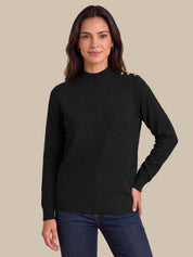 A woman wearing a black mock-neck knit sweater with gold button details on the shoulder, paired with dark blue jeans, against a solid beige background.