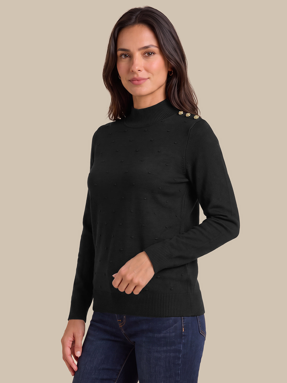 A woman modeling a black mock neck knit sweater with gold button details on the shoulders, paired with dark blue jeans, against a solid beige background.