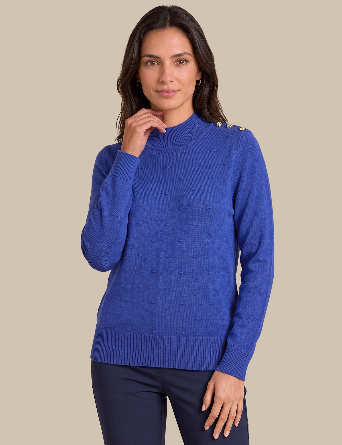 A woman modeling a royal blue knit sweater with textured bobble details and gold button accents on the shoulders, paired with dark navy pants, against a solid beige background.
