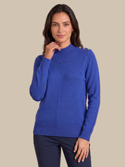 A woman modeling a royal blue knit sweater with textured bobble details and gold button accents on the shoulders, paired with dark navy pants, against a solid beige background.