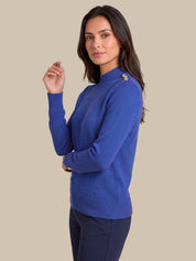 A woman modeling a royal blue mock-neck knit sweater with textured knit details and decorative gold-toned buttons on the shoulders, paired with dark navy trousers, against a solid beige background.