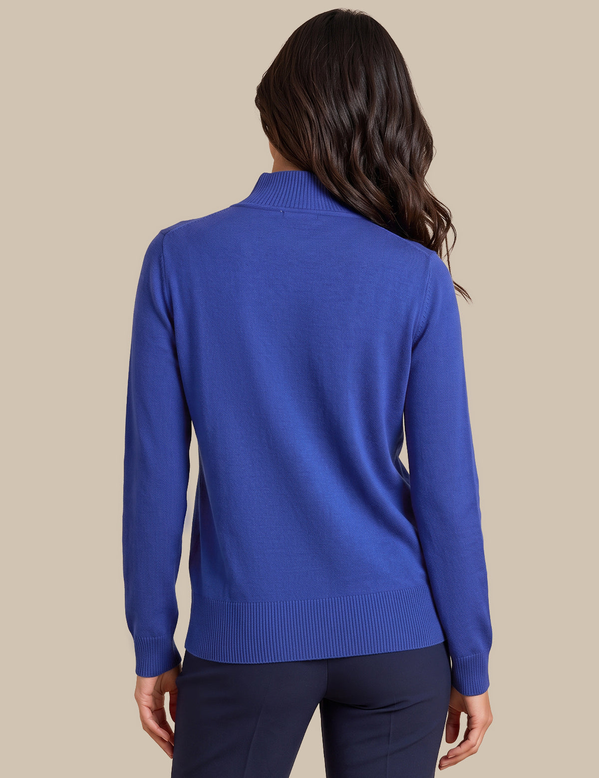 A woman shown from the back wearing a royal blue mock neck knit sweater and dark navy pants, set against a solid beige background.