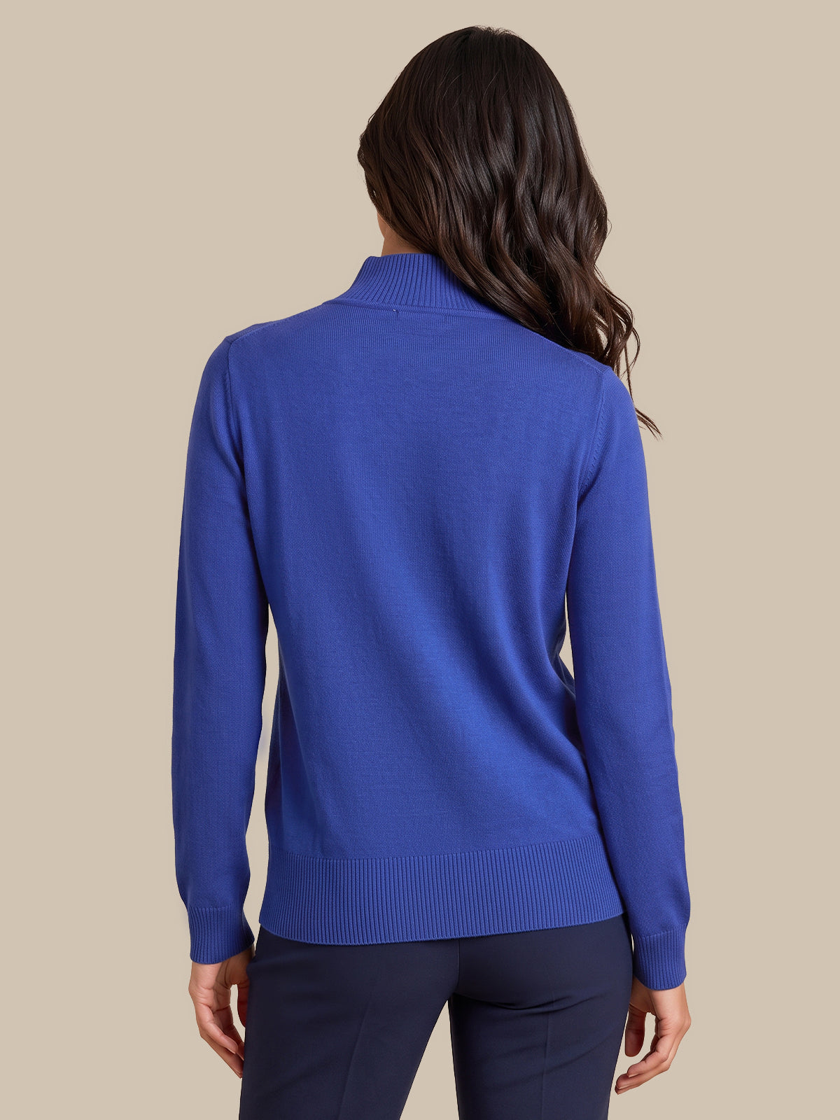 A woman shown from the back wearing a royal blue mock neck knit sweater and dark navy pants, set against a solid beige background.