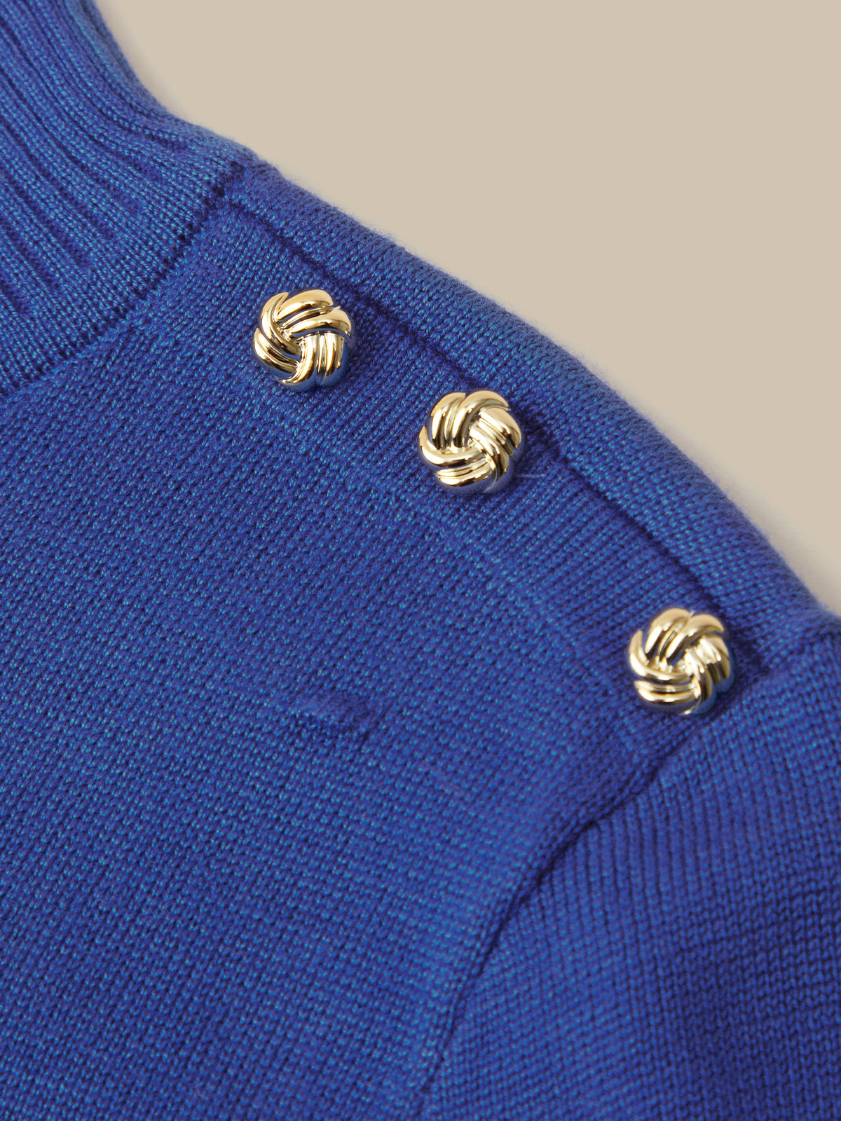 Close-up of a royal blue knit sweater featuring three decorative gold knotted buttons on the shoulder, set against a solid beige background.