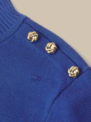 Close-up of a royal blue knit sweater featuring three decorative gold knotted buttons on the shoulder, set against a solid beige background.