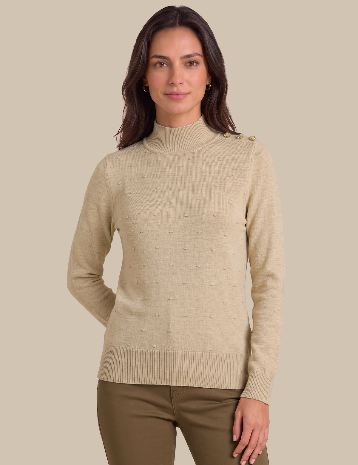 A woman wearing a beige ribbed mock neck sweater with subtle textured details and shoulder buttons, paired with brown pants, against a solid beige background.