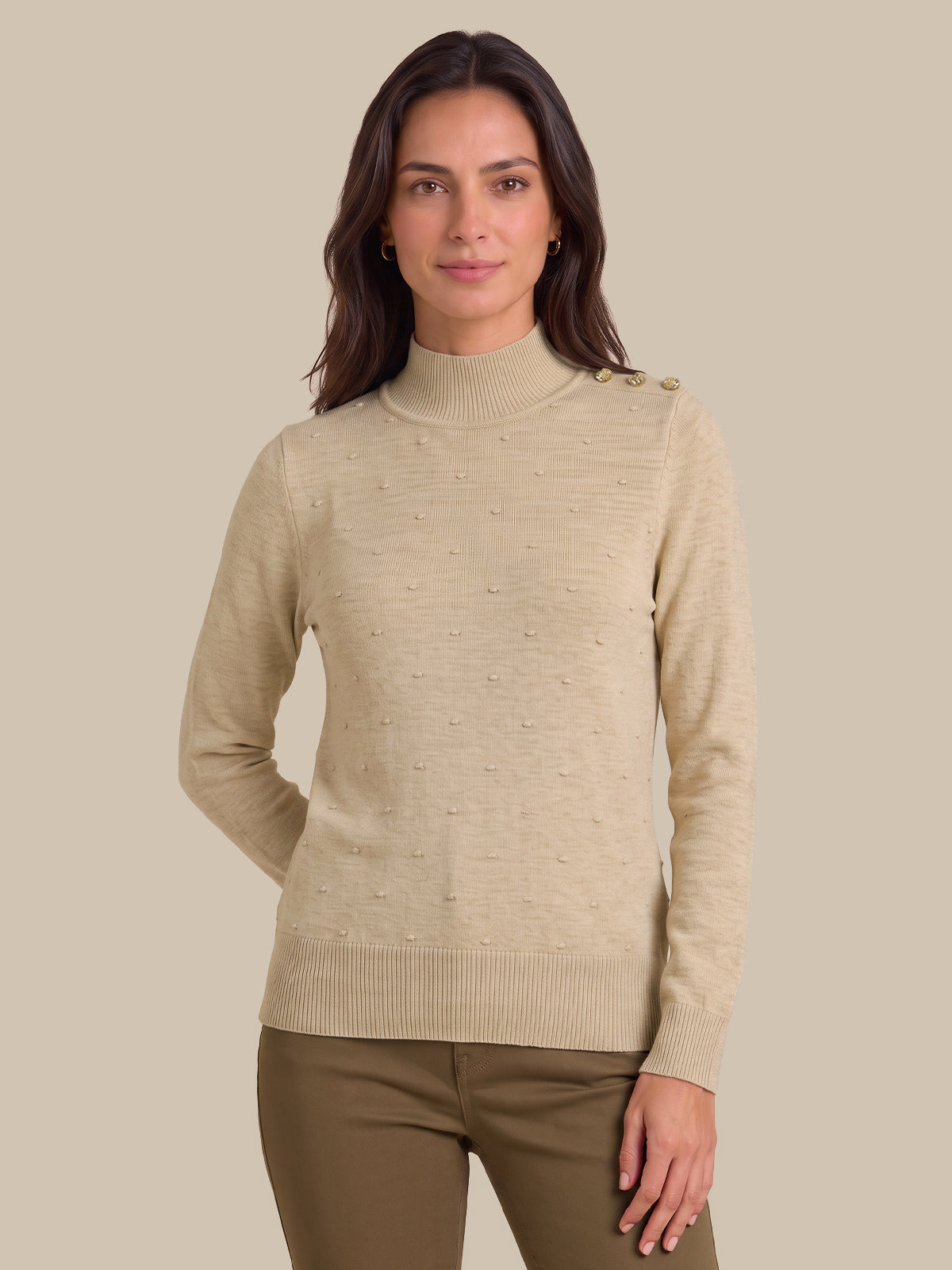 A woman wearing a beige ribbed mock neck sweater with subtle textured details and shoulder buttons, paired with brown pants, against a solid beige background.