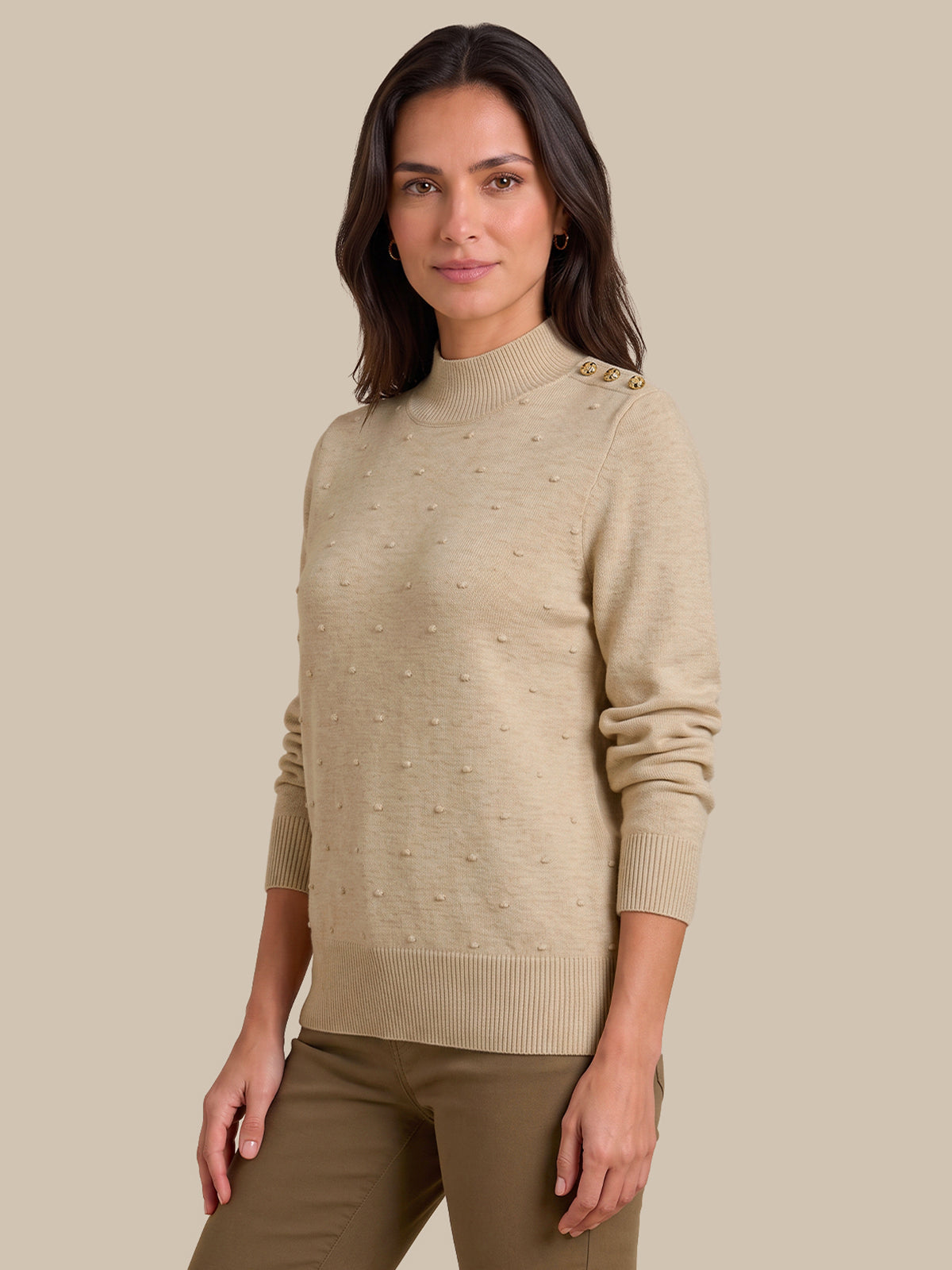 A woman modeling a beige knit sweater with textured pom-pom details and decorative buttons on the shoulders, paired with olive green pants, against a solid beige background.