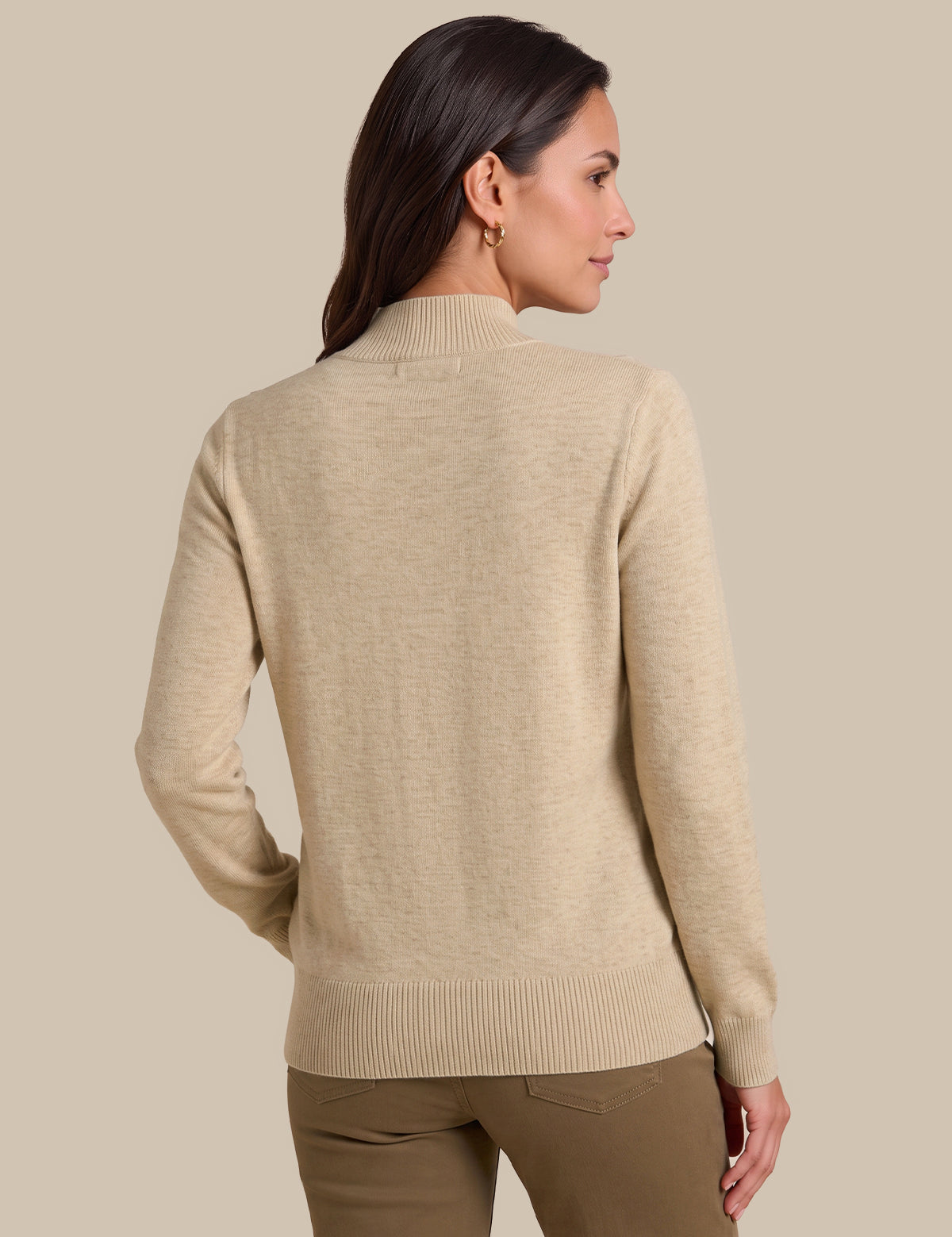 A woman shown from the back wearing a light beige knit sweater with a mock neck and ribbed hem and cuffs, paired with brown pants, against a solid beige background.