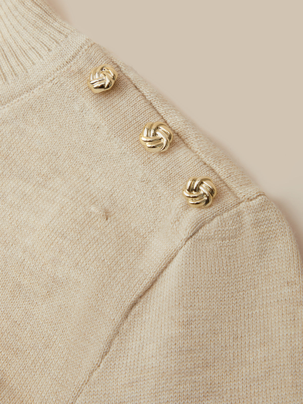 Close-up of a cream-colored knit sweater featuring three decorative gold knotted buttons along the shoulder seam.